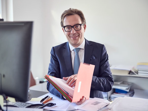 Ein geschäftsmännisch gekleideter Mann sitzt an einem Schreibtisch und hält einen bunten Aktendeckel in den Händen, während er lächelnd in die Kamera blickt. Auf dem Schreibtisch befinden sich Notizen, Schreibutensilien und ein Computer.
