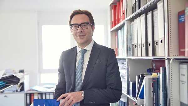 Ein Mann in einem Anzug und mit einer Brille steht in einem Büro und hält ein Buch in der Hand. Im Hintergrund sind Regale mit Aktenordnern und Büchern zu sehen.