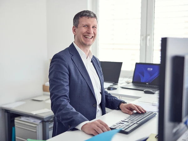 Ein lächelnder Mann sitzt an einem Schreibtisch, vor einem Computer, in einem modernen Büro mit mehreren Monitoren im Hintergrund.