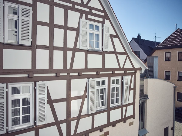 Ein Fachwerkhaus mit weißen Wänden und braunen Fachwerkbalken steht im Vordergrund, während im Hintergrund moderne Gebäude zu sehen sind. Der Himmel ist blau und die Sonne scheint.