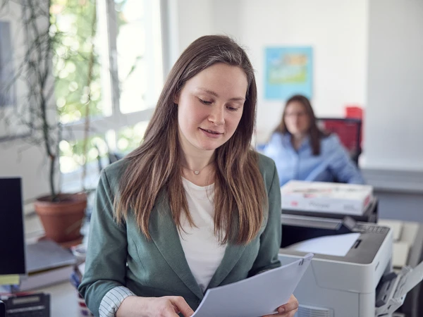Eine Frau mit langen, braunen Haaren trägt einen Blazer und liest Dokumente in einem modernen Büroraum, während eine weitere Person im Hintergrund an einem Schreibtisch sitzt.