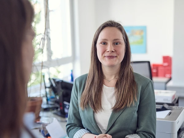 Eine Frau mit langen, braunen Haaren in einem grünen Blazer steht in einem modernen Büro und schaut freundlich in Richtung der Kamera. Im Hintergrund sind Büromaterialien und ein Drucker sichtbar.
