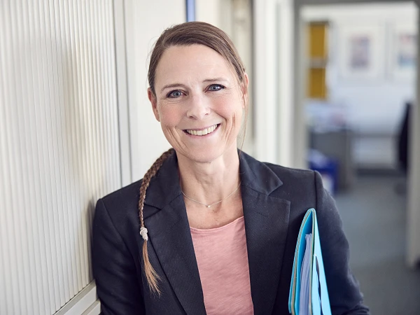 Eine lächelnde Frau mit langen, geflochtenen Haaren steht in einem Büro und hält einen Stapel Dokumente in der Hand. Sie trägt einen schwarzen Blazer über einem rosa Oberteil und steht an einer grauen Wand. Im Hintergrund sind verschwommen Büromöbel zu sehen.
