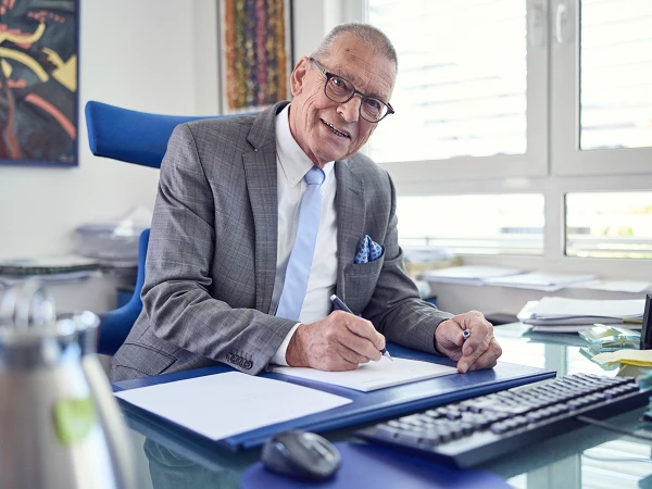 Ein älterer Mann in einem grauen Anzug mit einer Krawatte sitzt an einem Schreibtisch und schreibt auf einem Blatt Papier, während er lächelt. Im Hintergrund sind Dokumente und eine blühende Pflanze zu sehen.
