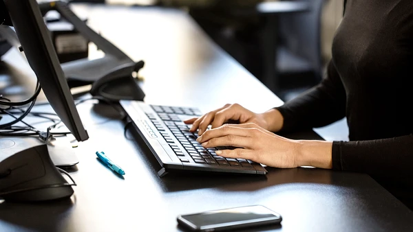 Eine Person tippt mit beiden Händen auf einer Tastatur, während sie an einem Schreibtisch sitzt. Auf dem Schreibtisch liegen ein Smartphone und ein blauer Stift.