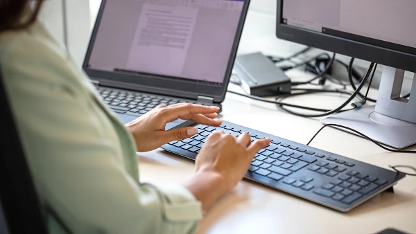 Eine Person tippt an einer Tastatur, während zwei Computerbildschirme auf einem Schreibtisch sichtbar sind, einer mit einem geöffneten Textdokument.