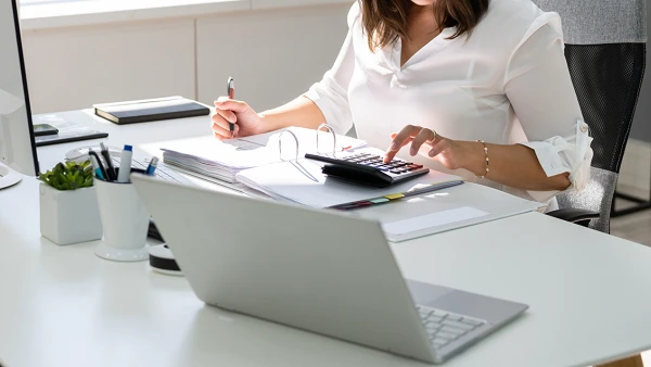 Eine Frau sitzt an einem modernen Schreibtisch in einem hellen Büro, während sie mit einem Taschenrechner arbeitet und Notizen auf einem Notizblock macht. Auf dem Tisch stehen mehrere Büromaterialien, ein Computerbildschirm und eine Laptop, während Sonnenlicht durch das Fenster strömt.