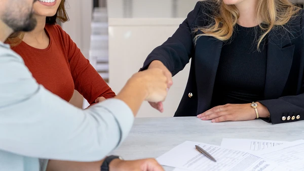 Eine blonde Frau in einem schwarzen Blazer lächelt, während sie einem Mann die Hand schüttelt. Neben ihm sitzt eine Frau mit dunklen, lockigen Haaren und einer roten Bluse, die ebenfalls lächelt. Auf dem Tisch liegen Dokumente und ein Stift.