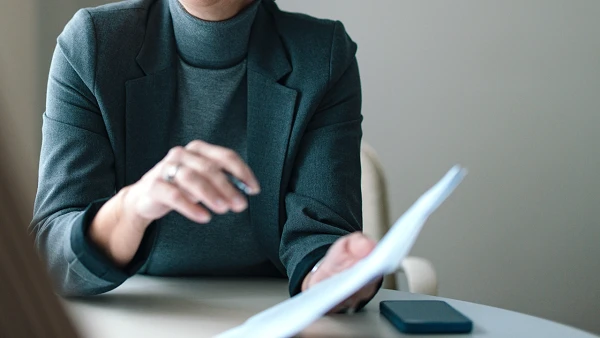 Eine lächelnde Frau mit kurzen grauen Haaren und Brille sitzt in einem modernen Büro und hält Unterlagen in der Hand, während sie ein Gespräch führt. Im Hintergrund sind verschwommen eine weitere Person sowie einige Wandbilder zu sehen.