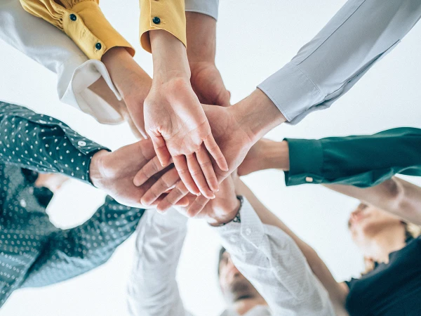 Mehrere Hände verschiedener Personen sind in einem Kreis zusammengelegt, während sich die Menschen um die Hände gruppieren. Die Hände sind in unterschiedlichen Hauttönen und Kleidungsstilen, was Vielfalt und Zusammenarbeit symbolisiert.