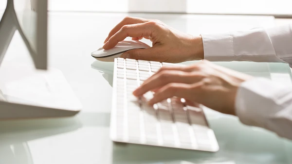 Eine Hand verwendet eine Computermaus, während die andere Hand auf einer weißen Tastatur tippt. Im Hintergrund ist ein Bildschirm auf einem Tisch zu sehen.
