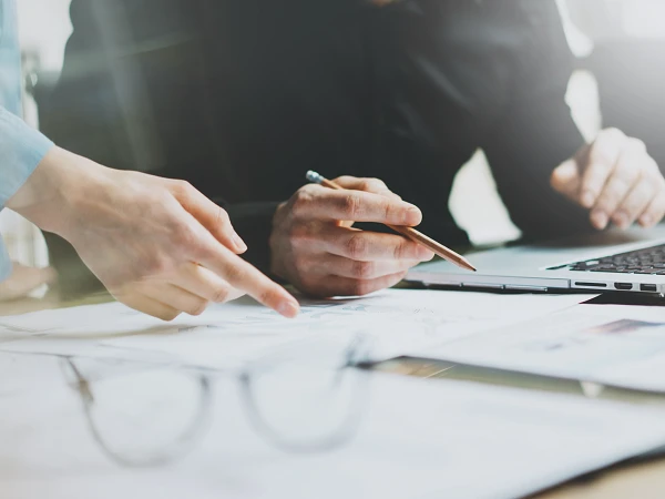 Zwei Personen arbeiten gemeinsam an einem Tisch, während eine Hand auf ein Papier zeigt und die andere einen Stift hält; ein Laptop ist geöffnet und leicht sichtbar, sowie Brille und weitere Papiere liegen davor.