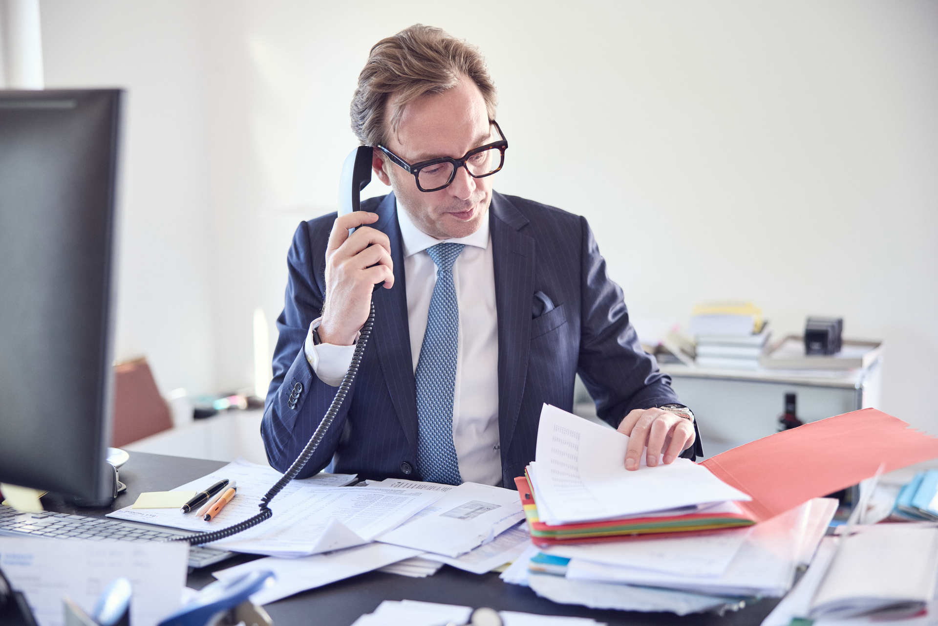 Ein Geschäftsmann mit Brille spricht am Telefon, während er mehrere Dokumente auf seinem Schreibtisch durchblättert, der unordentlich mit Papieren und Büromaterialien bedeckt ist.