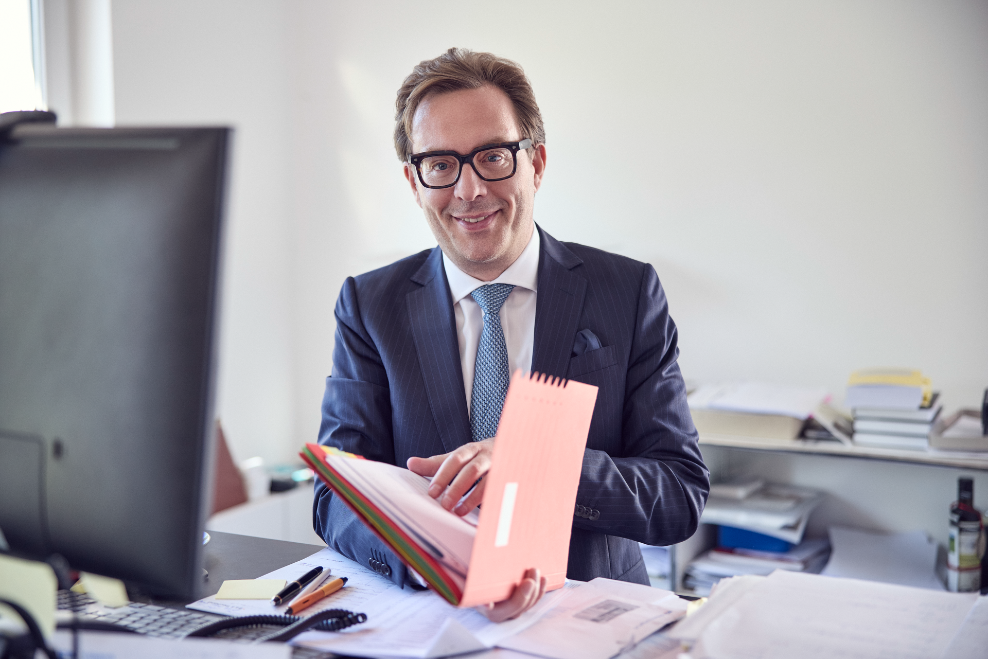 Ein geschäftsmännisch gekleideter Mann sitzt an einem Schreibtisch und hält einen bunten Aktendeckel in den Händen, während er lächelnd in die Kamera blickt. Auf dem Schreibtisch befinden sich Notizen, Schreibutensilien und ein Computer.