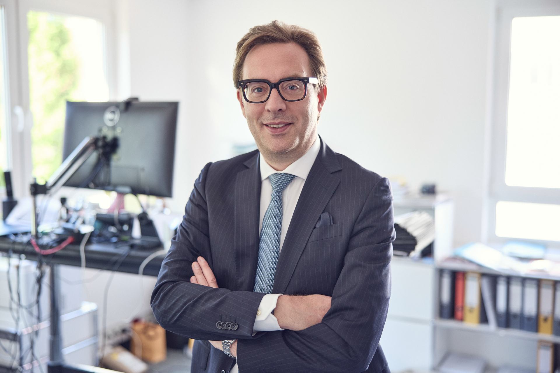 Ein Mann in einem dunkelblauen Anzug mit Krawatte steht mit verschränkten Armen in einem modernen Büro, das einen Computer und Bürobedarf im Hintergrund zeigt.
