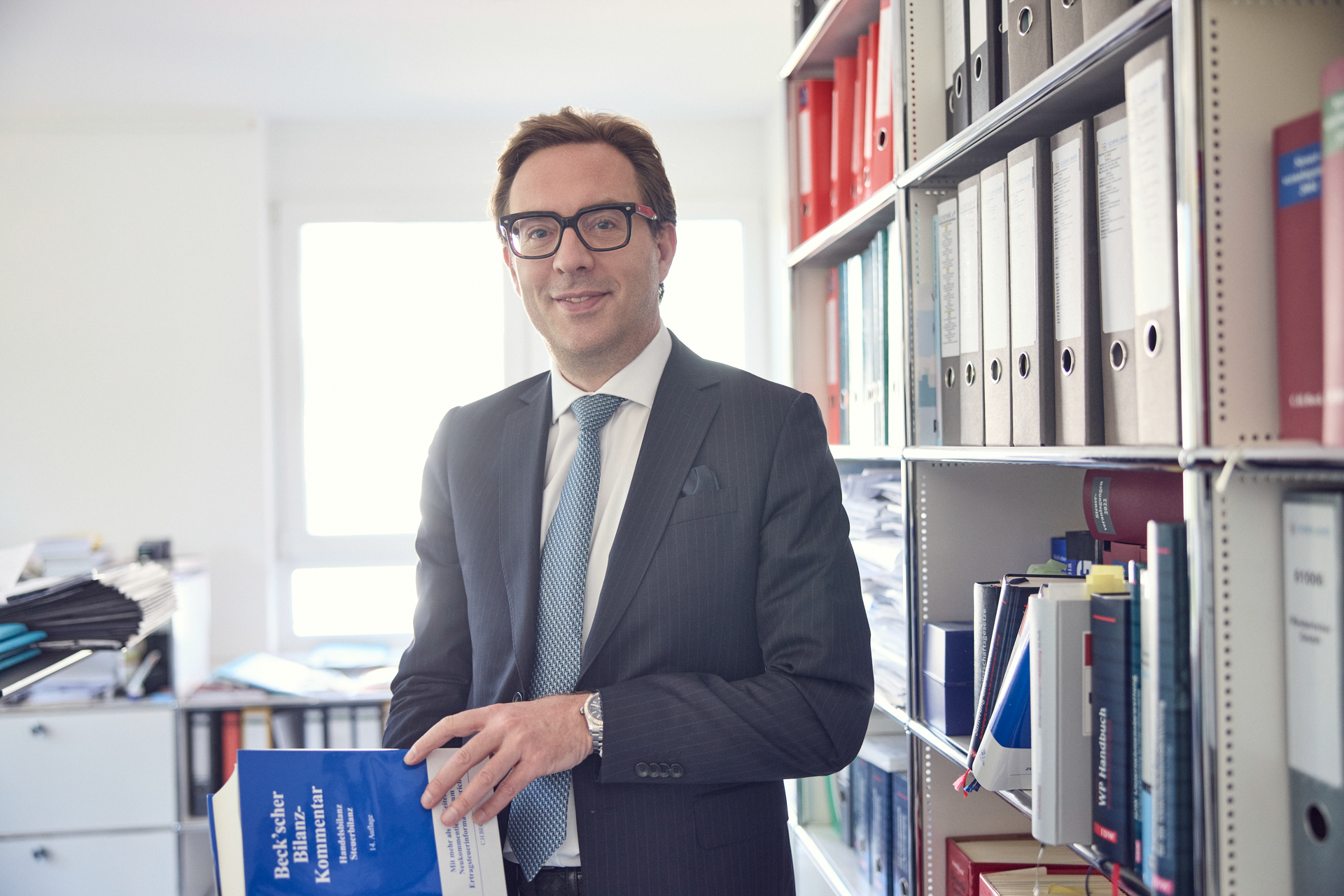 Ein Mann in einem Anzug und mit einer Brille steht in einem Büro und hält ein Buch in der Hand. Im Hintergrund sind Regale mit Aktenordnern und Büchern zu sehen.