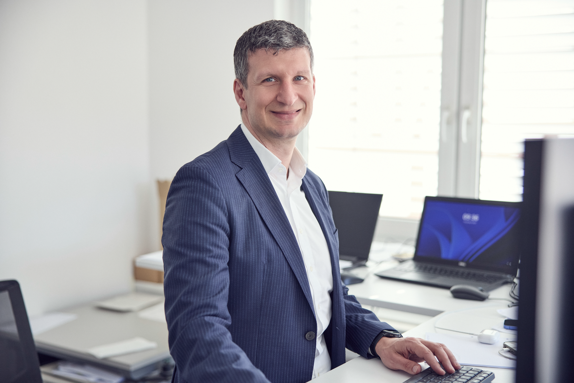 Ein Mann in einem blauen Anzug sitzt an einem Schreibtisch in einem modernen Büro mit mehreren Laptops im Hintergrund und lächelt in die Kamera.