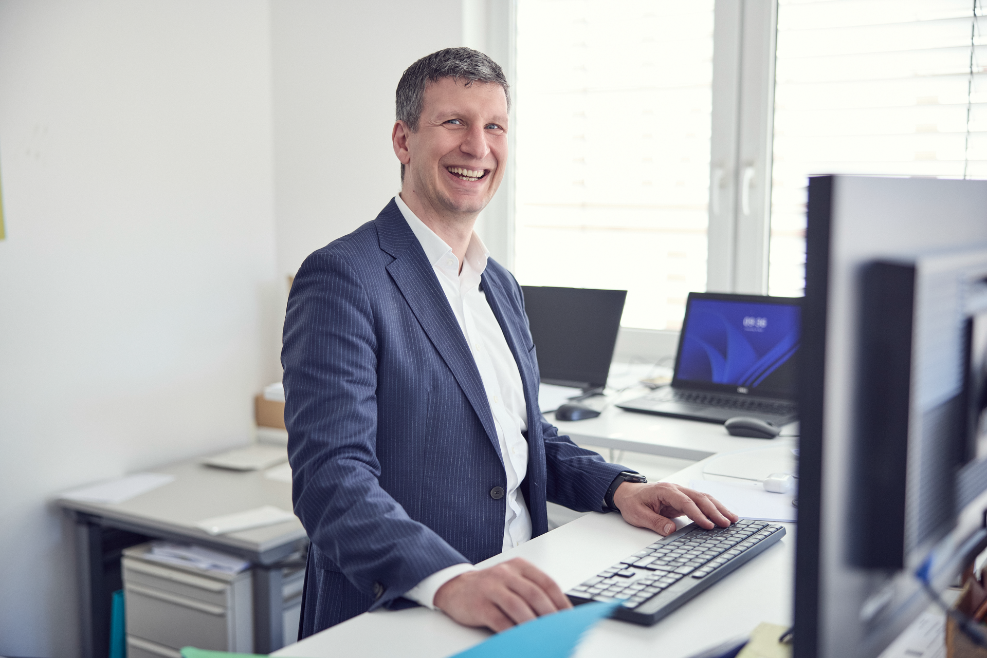Ein lächelnder Mann sitzt an einem Schreibtisch, vor einem Computer, in einem modernen Büro mit mehreren Monitoren im Hintergrund.