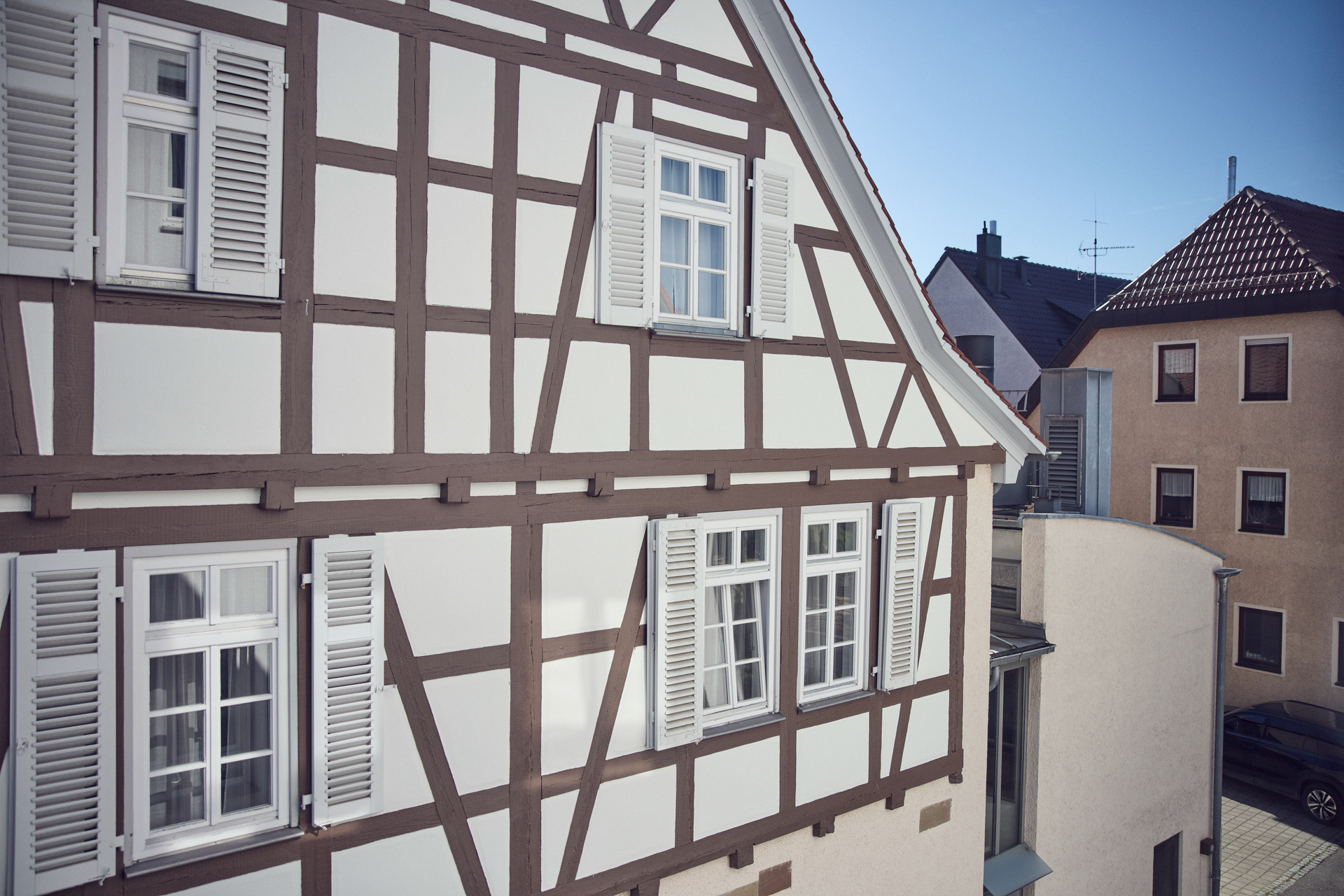Ein Fachwerkhaus mit weißen Wänden und braunen Fachwerkbalken steht im Vordergrund, während im Hintergrund moderne Gebäude zu sehen sind. Der Himmel ist blau und die Sonne scheint.
