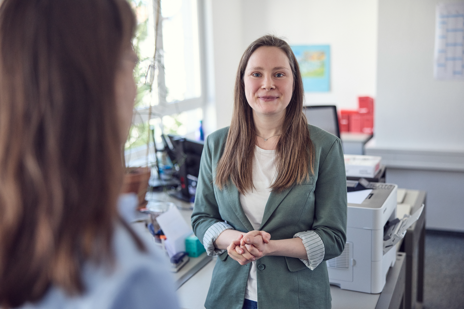 Eine Frau mit langen, braunen Haaren in einem grünen Blazer steht in einem modernen Büro und schaut freundlich in Richtung der Kamera. Im Hintergrund sind Büromaterialien und ein Drucker sichtbar.