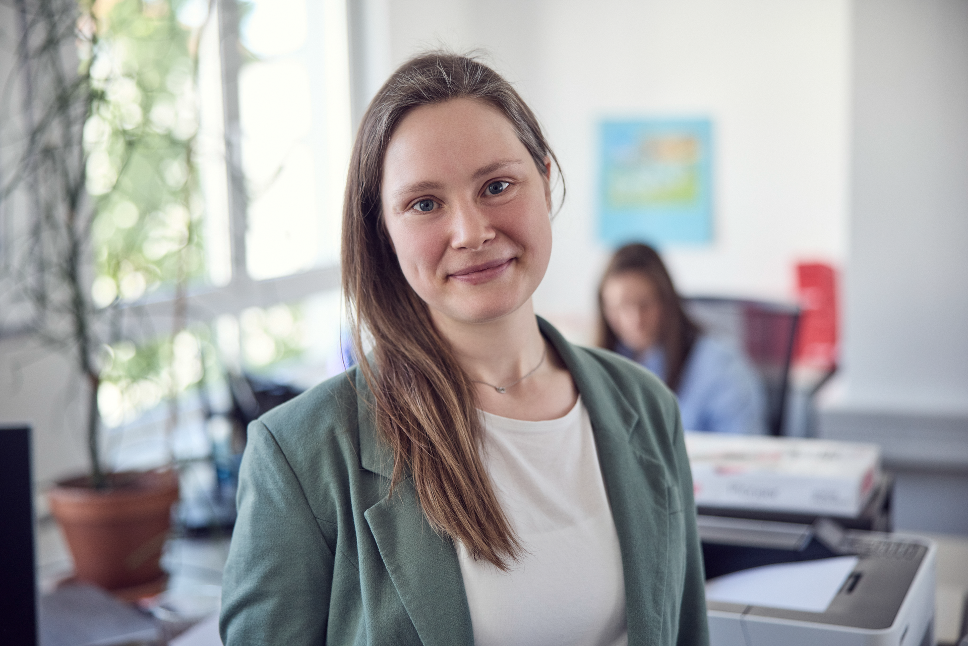 Eine junge Frau mit langen, bräunlichen Haaren trägt eine grüne Jacke und lächelt freundlich in die Kamera, während sie in einem modernen Büro steht. Im Hintergrund ist eine weitere Person an einem Schreibtisch zu sehen, die an einem Computer arbeitet.