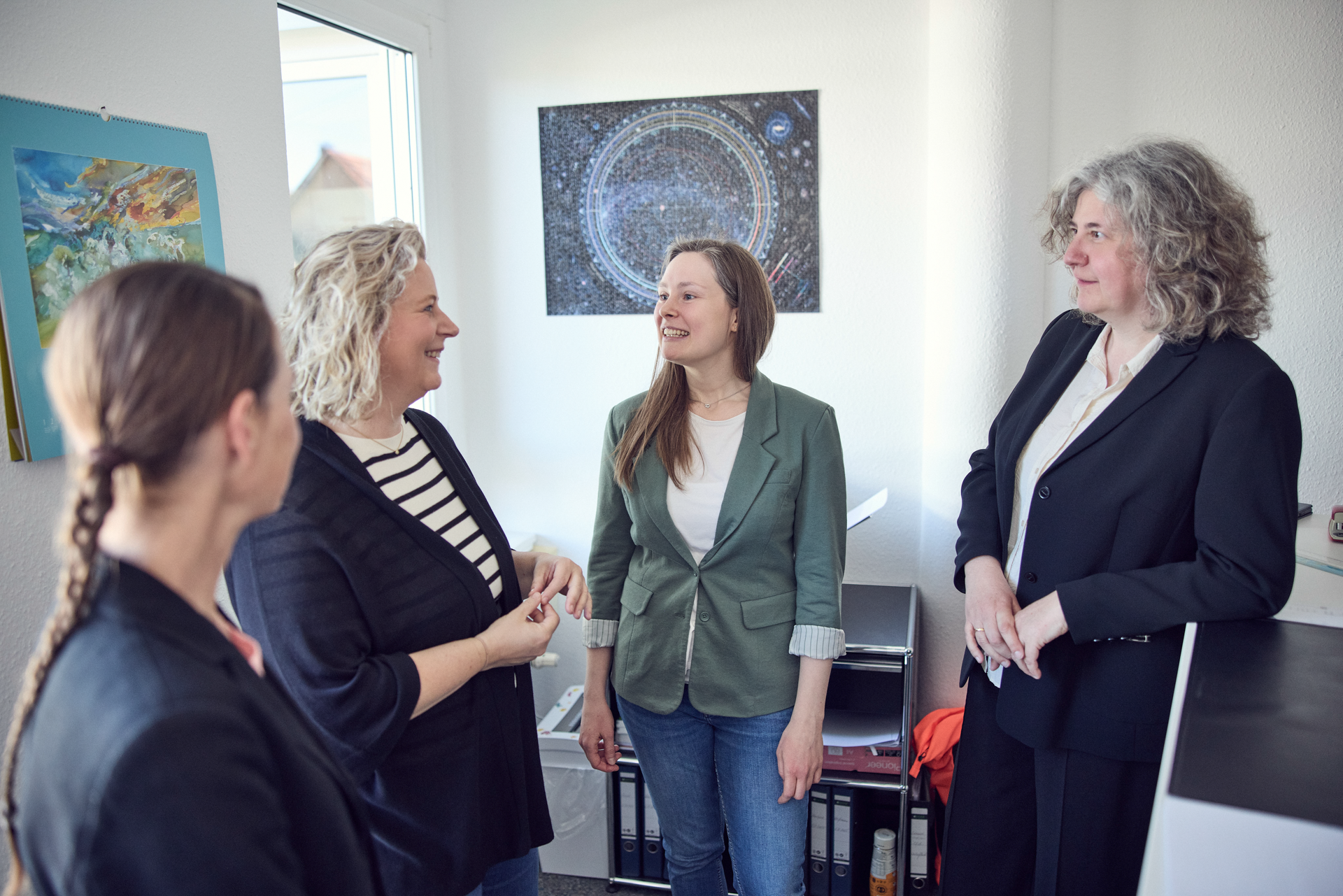 Vier Frauen stehen in einem Büro und unterhalten sich. Eine der Frauen trägt einen grünen Blazer, eine weitere hat lockiges Haar. Im Hintergrund ist ein großes Kunstwerk an der Wand zu sehen.