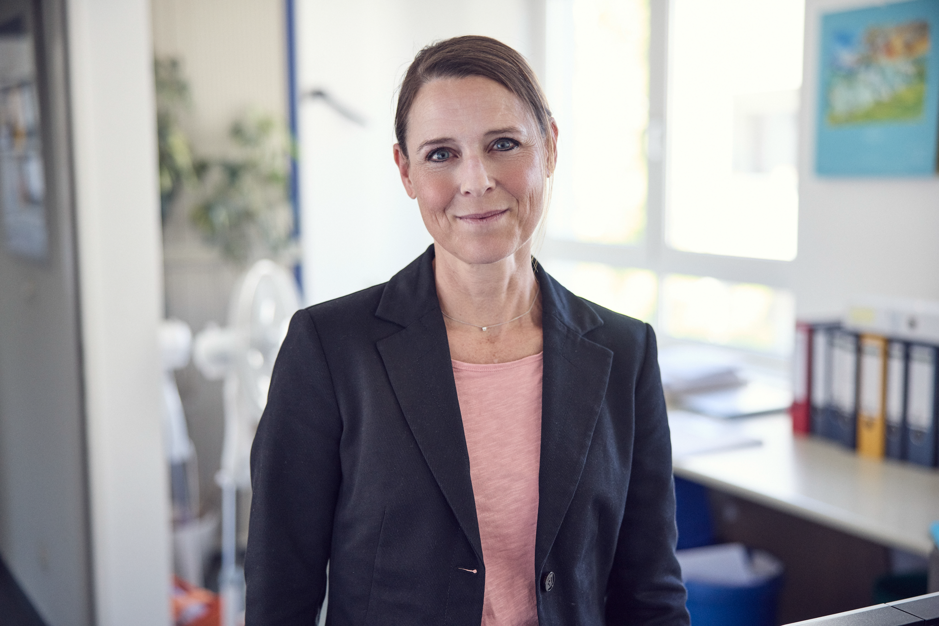 Eine Frau mit langen, braunen Haaren trägt einen schwarzen Blazer und ein rosa Oberteil. Sie steht in einem modernen Büro mit hellen Wänden und Schreibtischunterlagen im Hintergrund. In ihrem Gesichtsausdruck zeigt sich ein freundliches Lächeln.