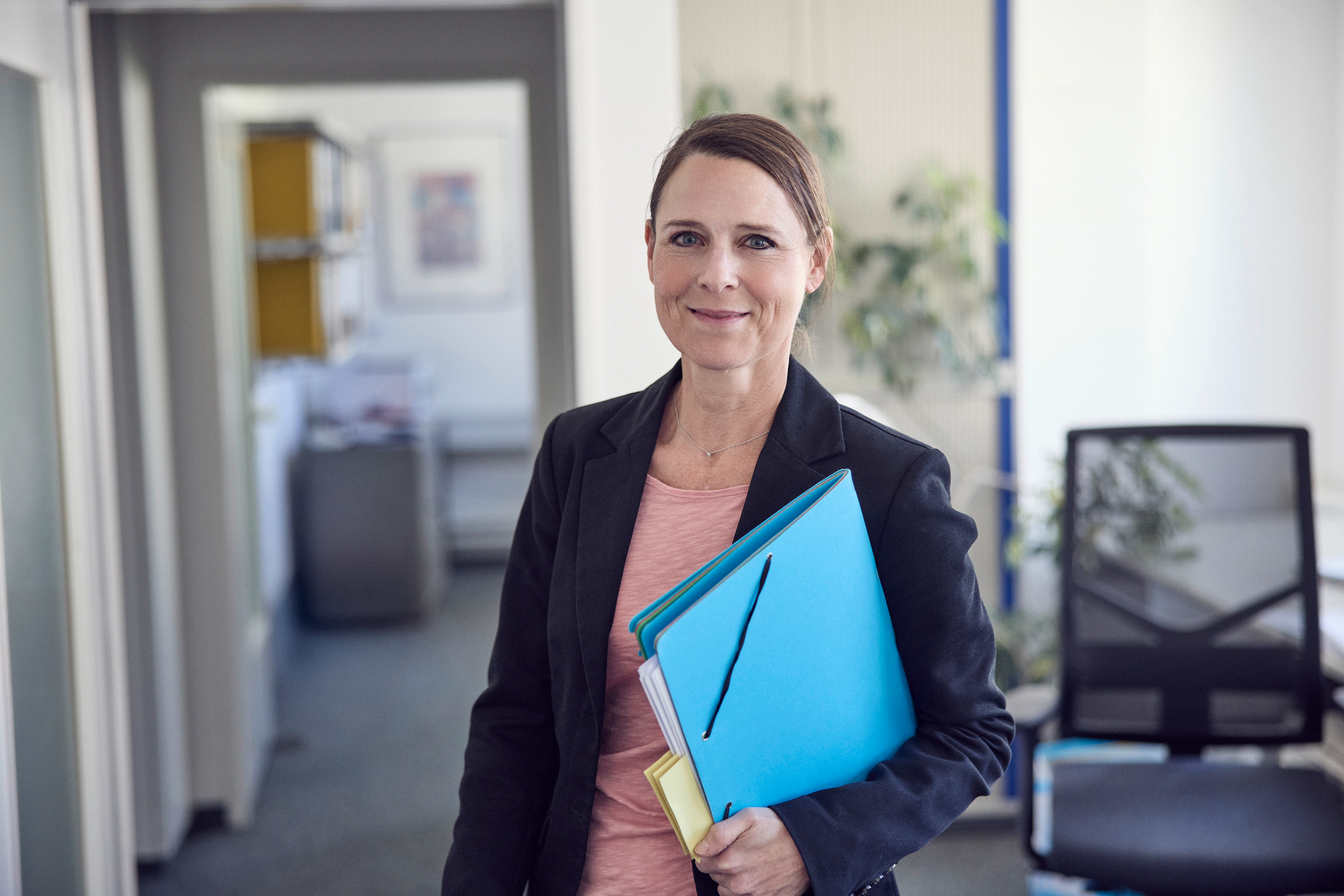 Eine Frau mit braunen Haaren, die in einem blauen Blazer gekleidet ist, steht in einem Büroumfeld und hält mehrere farbige Ordner in ihren Händen. Im Hintergrund sind Büromöbel und Pflanzen sichtbar.