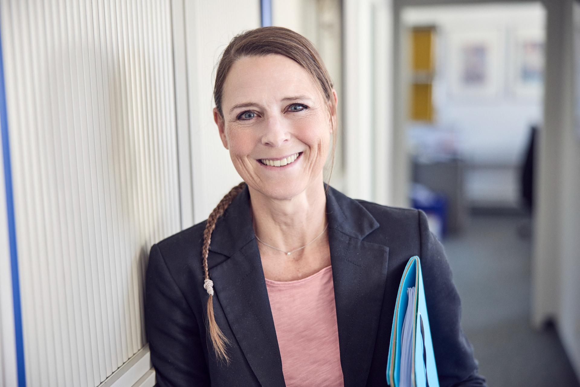 Eine lächelnde Frau mit langen, geflochtenen Haaren steht in einem Büro und hält einen Stapel Dokumente in der Hand. Sie trägt einen schwarzen Blazer über einem rosa Oberteil und steht an einer grauen Wand. Im Hintergrund sind verschwommen Büromöbel zu sehen.