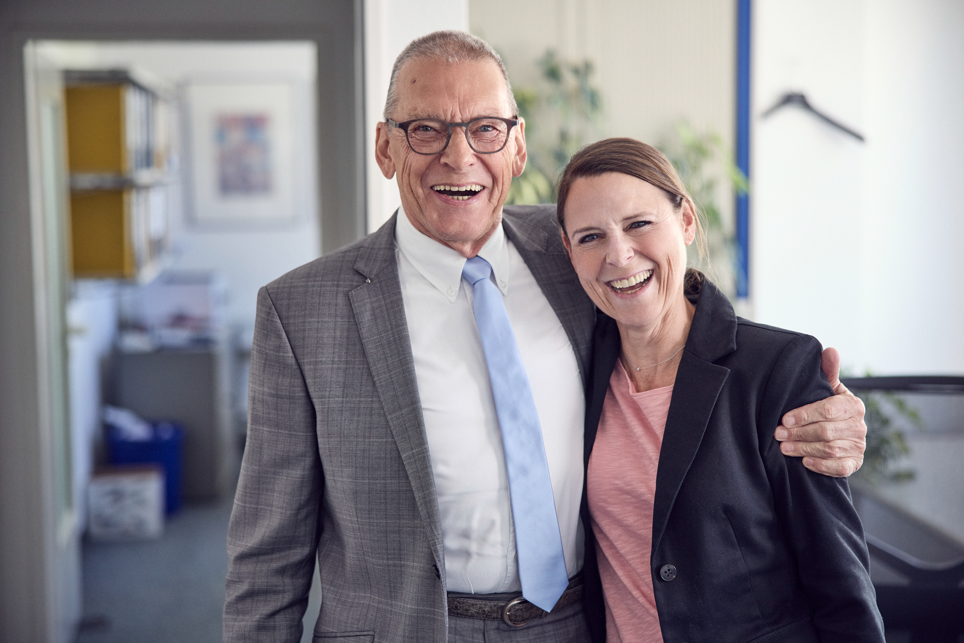 Ein älterer Mann mit Brille in einem grauen Anzug und eine lächelnde Frau in einem schwarzen Blazer stehen eng zusammen und posieren fröhlich für das Foto in einem Büro.