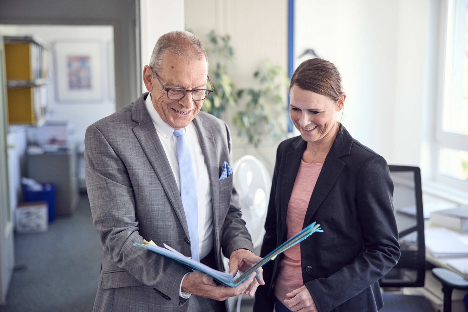 Ein älterer Mann in einem grauen Anzug und eine Frau in einem schwarzen Blazer stehen in einem Büro, während sie gemeinsam in einen Ordner mit Unterlagen schauen und lächeln.