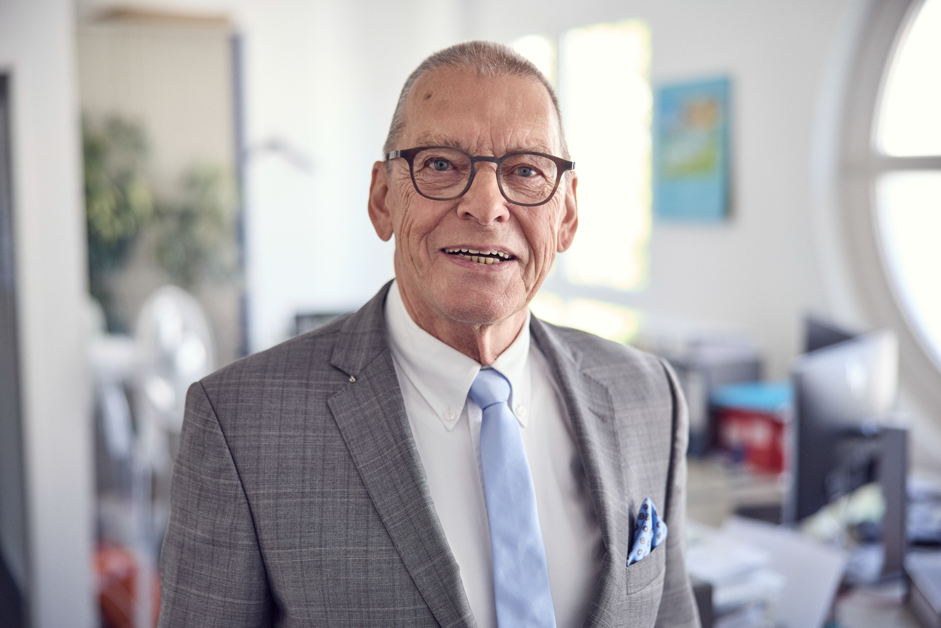 Ein älterer Mann mit Brille trägt einen grauen Anzug und ein hellblaues Hemd mit Krawatte, während er in einem modernen Büro lächelt. Im Hintergrund sind Schreibtische und Computer zu sehen.