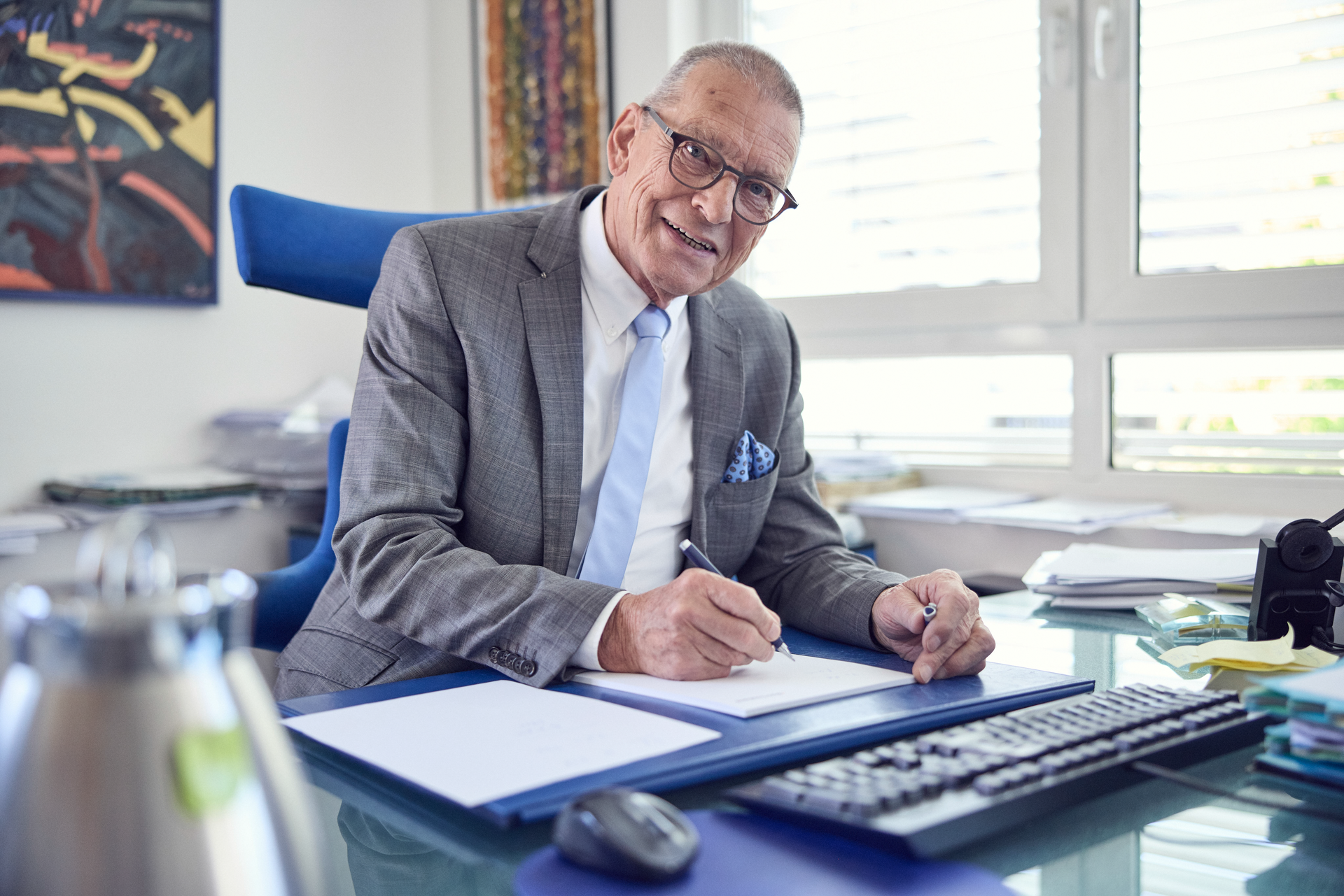 Ein älterer Mann in einem grauen Anzug mit einer Krawatte sitzt an einem Schreibtisch und schreibt auf einem Blatt Papier, während er lächelt. Im Hintergrund sind Dokumente und eine blühende Pflanze zu sehen.