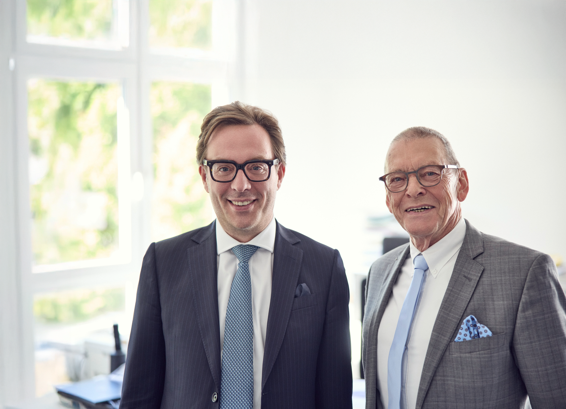 Zwei Männer in Anzügen posieren lächelnd in einem hellen Büroumfeld. Der jüngere Mann trägt eine Brille und hat eine hellblaue Krawatte, während der ältere Mann eine graue Jacke und eine hellblaue Einstecktuch trägt. Im Hintergrund sind Büromöbel und helle Fenster zu sehen.