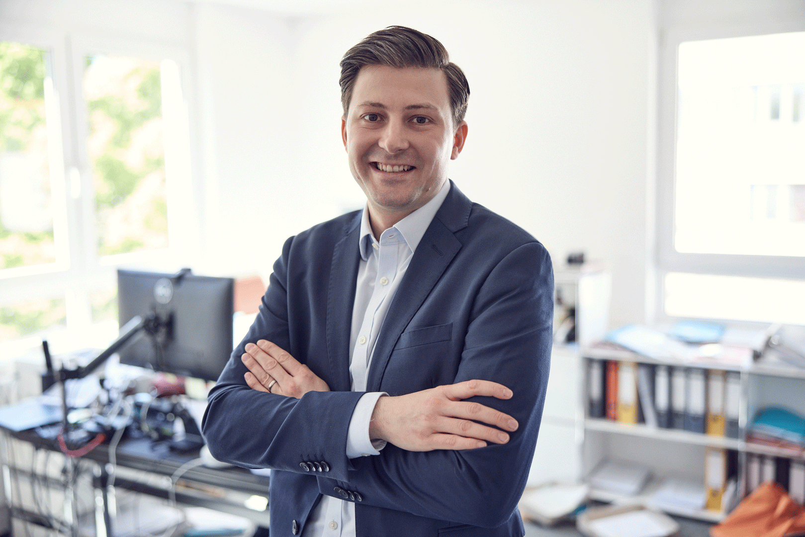 Ein lächelnder Mann in einem modernen Büro steht mit verschränkten Armen vor einem Schreibtisch, auf dem ein Computer und verschiedene organisatorische Materialien zu sehen sind.