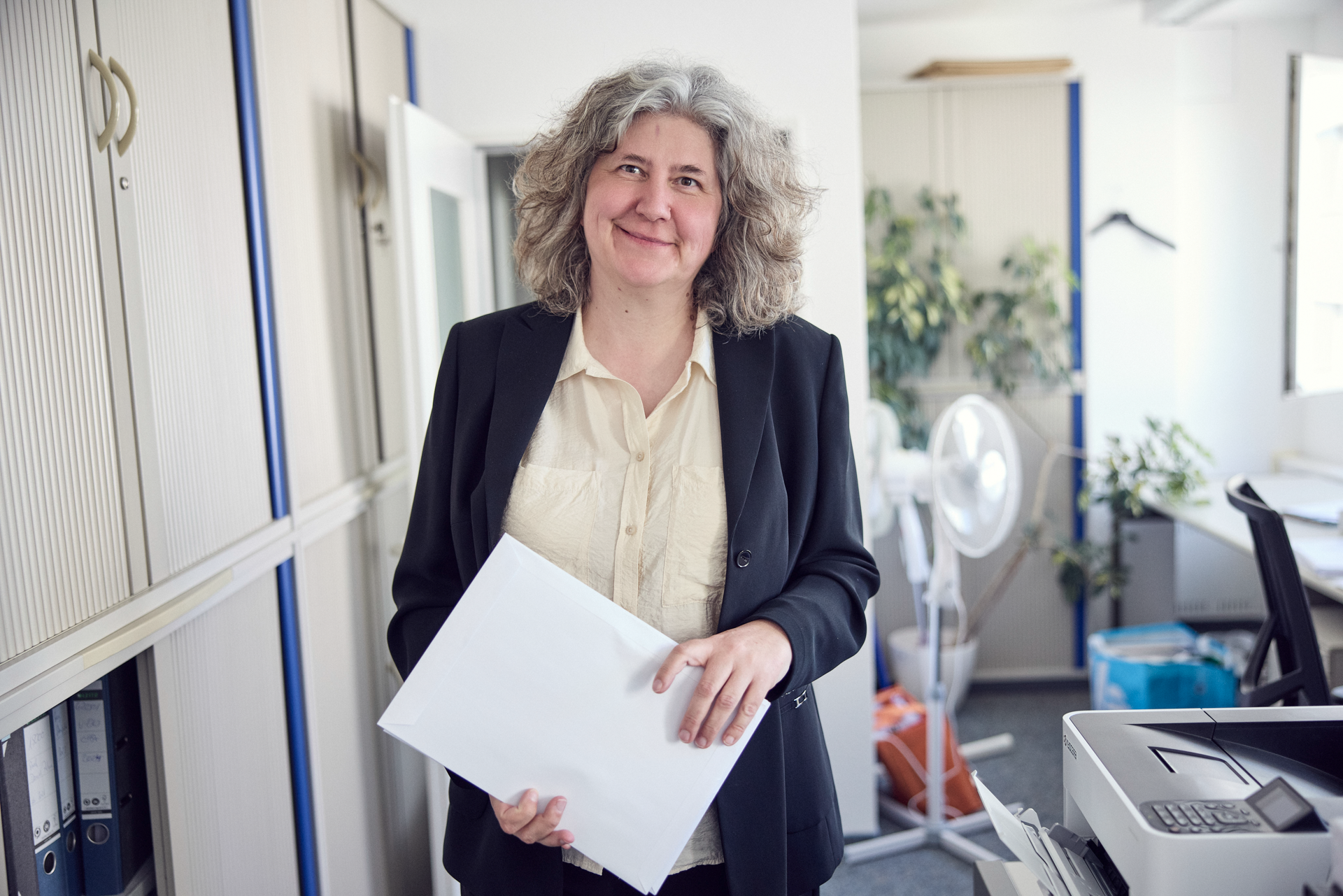 Eine Frau mit lockigem grauem Haar steht in einem Büroumfeld und hält ein Blatt Papier in der Hand, während sie lächelt. Im Hintergrund sind Schreibtische und Pflanzen zu sehen.