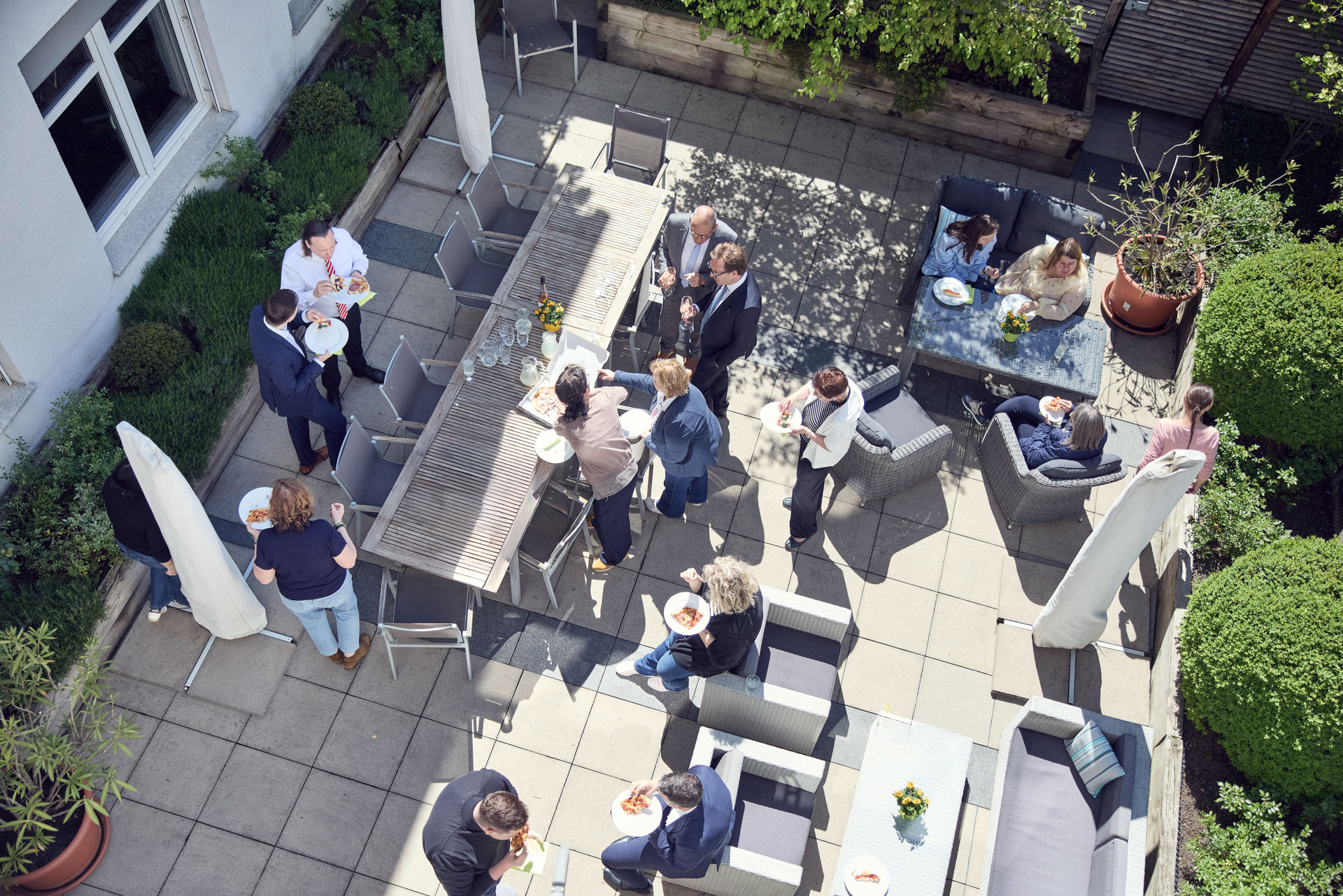 Eine Gruppe von Menschen steht und sitzt im Freien in einem Garten mit gepflastertem Boden, umgeben von Pflanzen. Einige Personen unterhalten sich an einem langen Tisch, während andere an Sofas in der Nähe essen und trinken. Es ist sonnig und die Atmosphäre wirkt entspannt und gesellig.