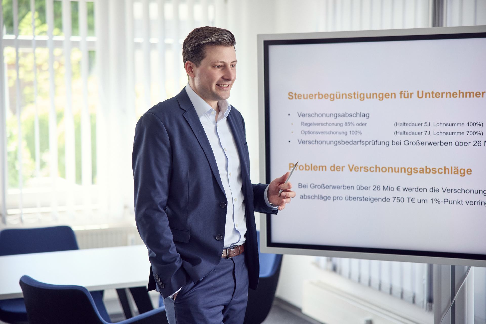 Ein Mann in einem Anzug steht neben einem Bildschirm, auf dem Text über Steuervergünstigungen für Unternehmen angezeigt wird. Er zeigt auf den Bildschirm und diskutiert die Thematik. Im Hintergrund sind helle Fenster und Büroeinrichtungen zu sehen.