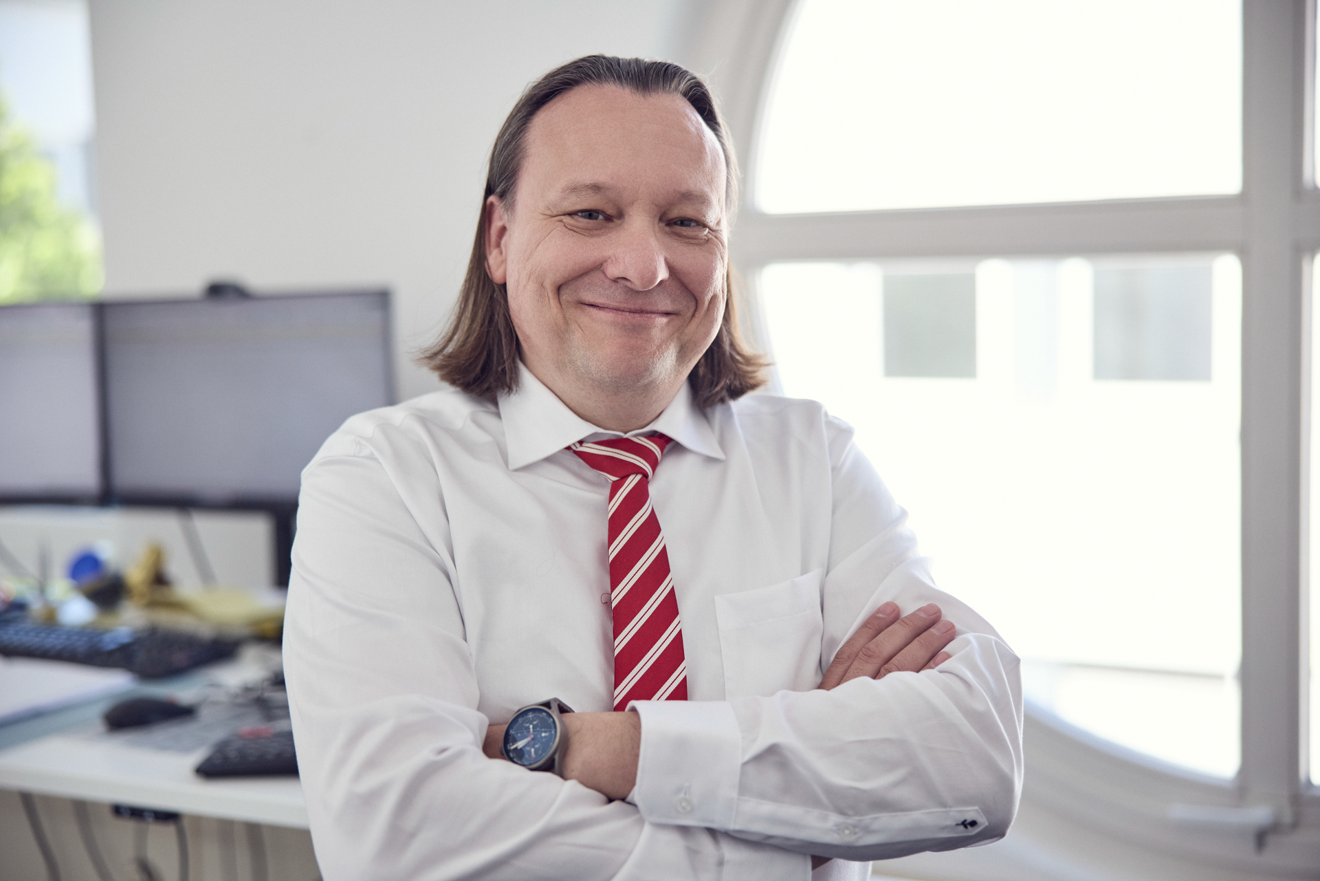 Ein Mann mit langen, dunkelblonden Haaren und einem freundlichen Lächeln steht mit übereinander geschlagenen Armen vor einem hellen Bürofenster. Er trägt ein weißes Hemd und eine rot-gestreifte Krawatte. Im Hintergrund sind Computer und Büromaterialien sichtbar.