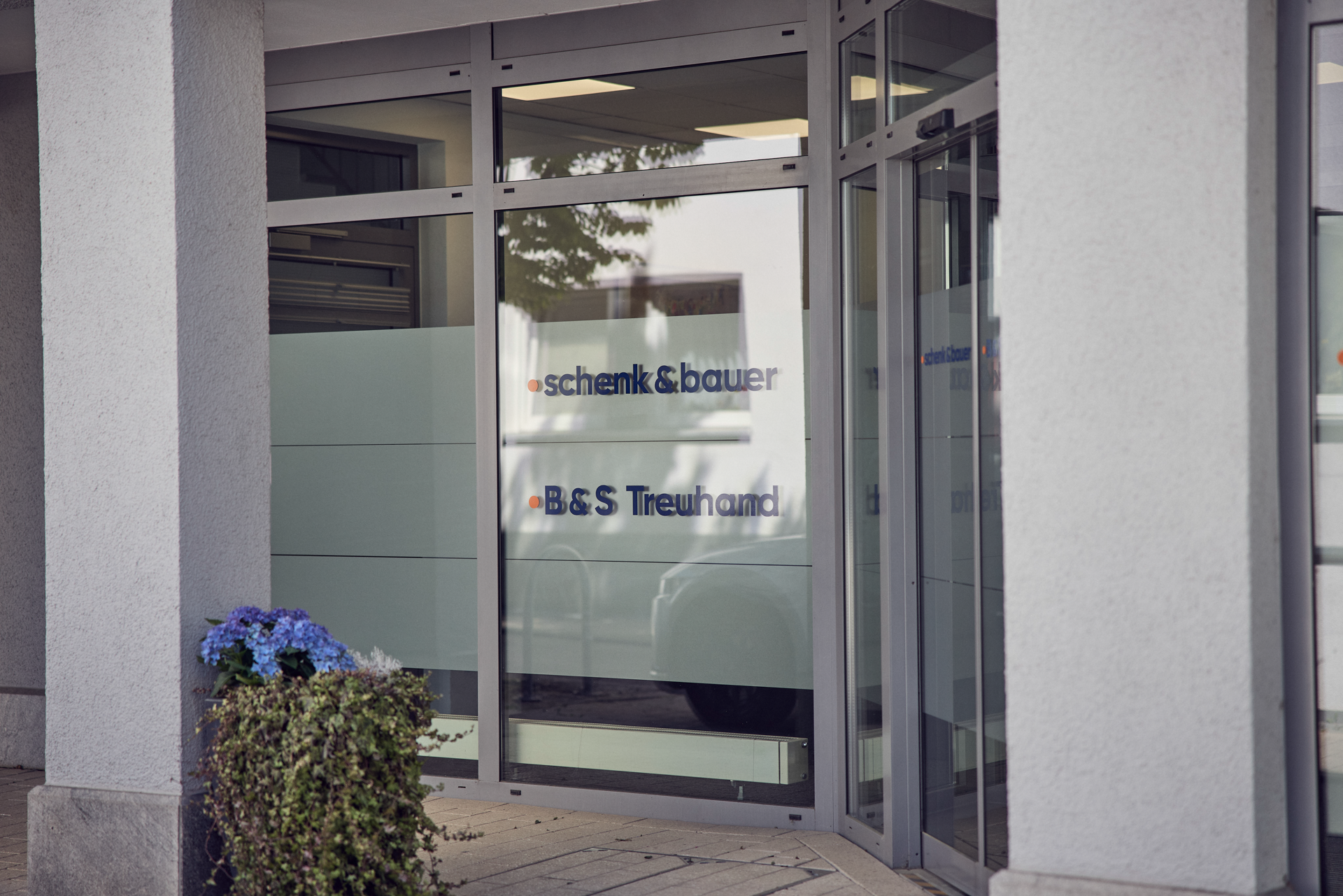 Das Bild zeigt den Eingangsbereich eines Geschäfts mit großen Glasfenstern, auf denen die Namen "schenk & bauer" und "B&S Treuhand" zu sehen sind. Vor dem Eingang steht ein Topf mit blauen Blumen.