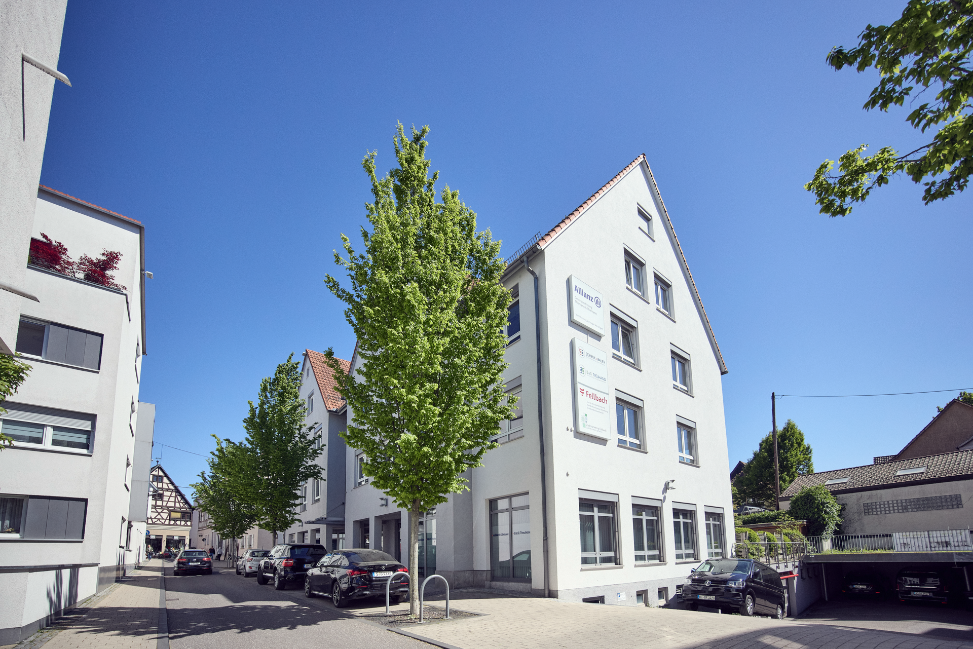 Ein dreieckiges, weißes Gebäude mit mehreren Fenstern und einem Schild an der Fassade steht an einer ruhigen Straße, flankiert von mehreren hohen Bäumen und geparkten Autos, während der Himmel strahlend blau ist.