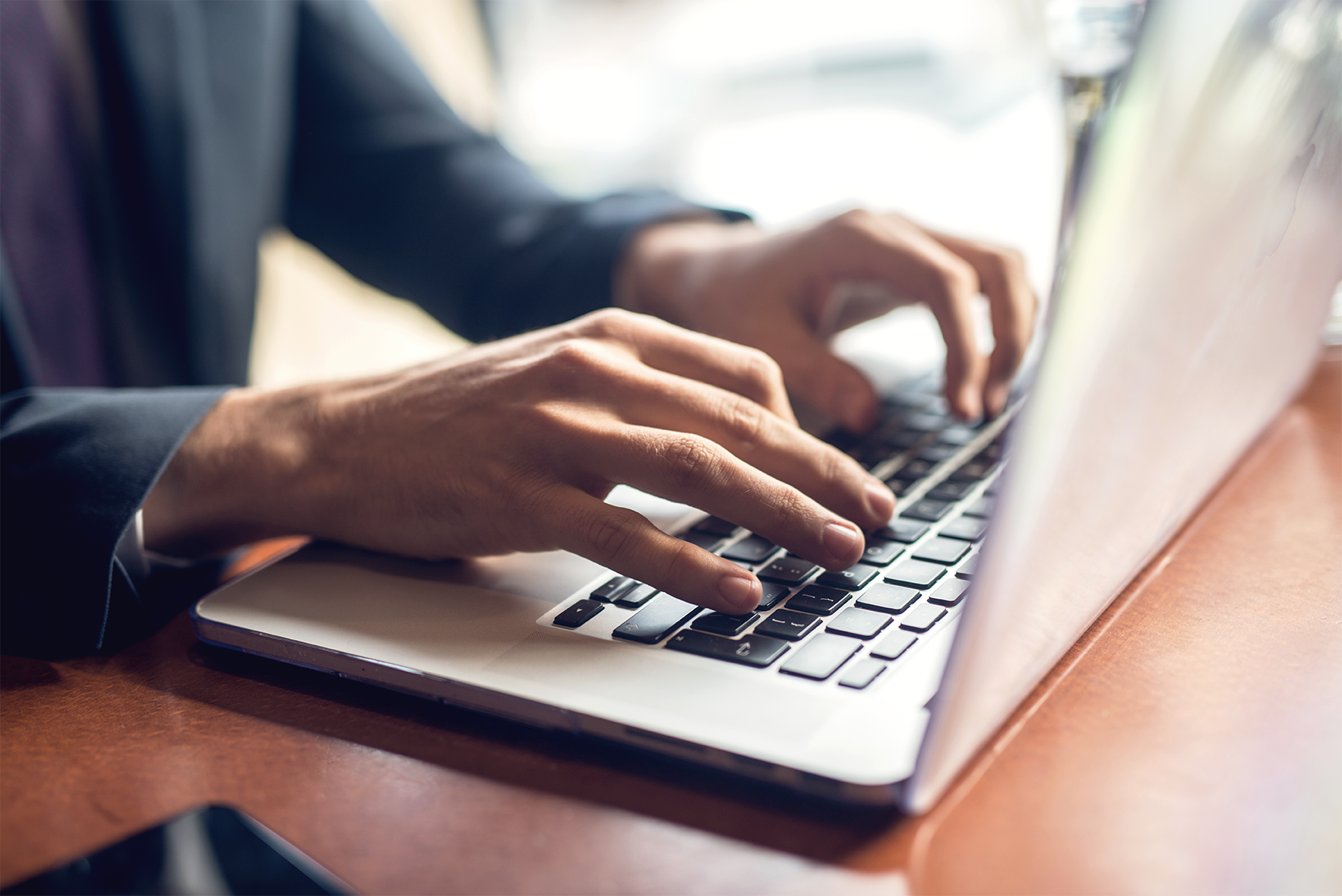 Eine Person in einem Anzug tippt auf einer Laptop-Tastatur, während sie an einem Tisch sitzt. Im Hintergrund sind verschwommene Gegenstände zu erkennen.