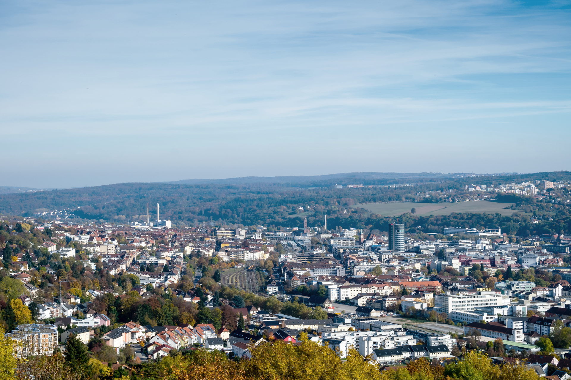 Blick auf eine Stadtlandschaft mit einer Mischung aus modernen und traditionellen Gebäuden, umgeben von sanften Hügeln und Bäumen im Herbst. Der Himmel ist klar und blau.