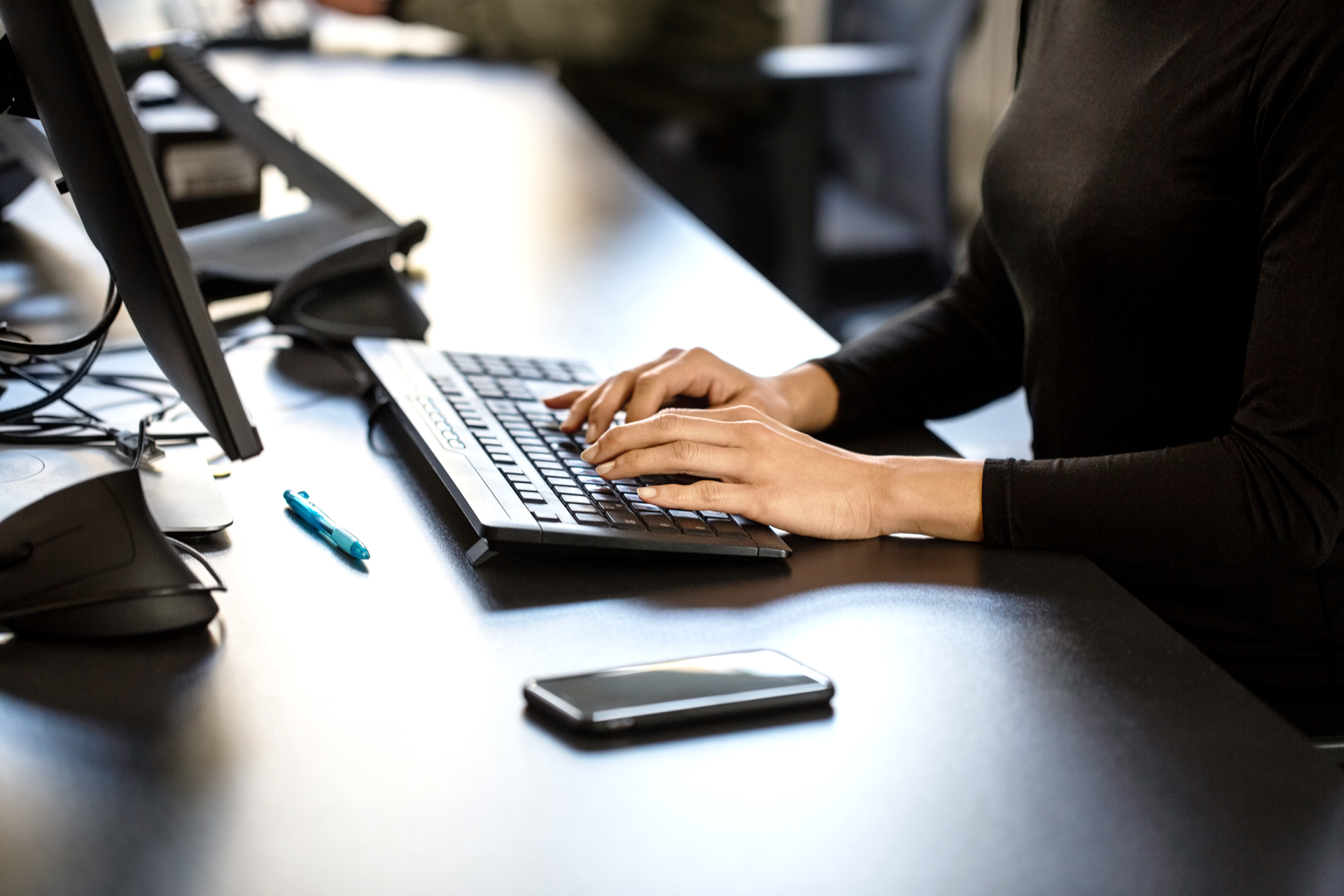 Eine Person tippt mit beiden Händen auf einer Tastatur, während sie an einem Schreibtisch sitzt. Auf dem Schreibtisch liegen ein Smartphone und ein blauer Stift.