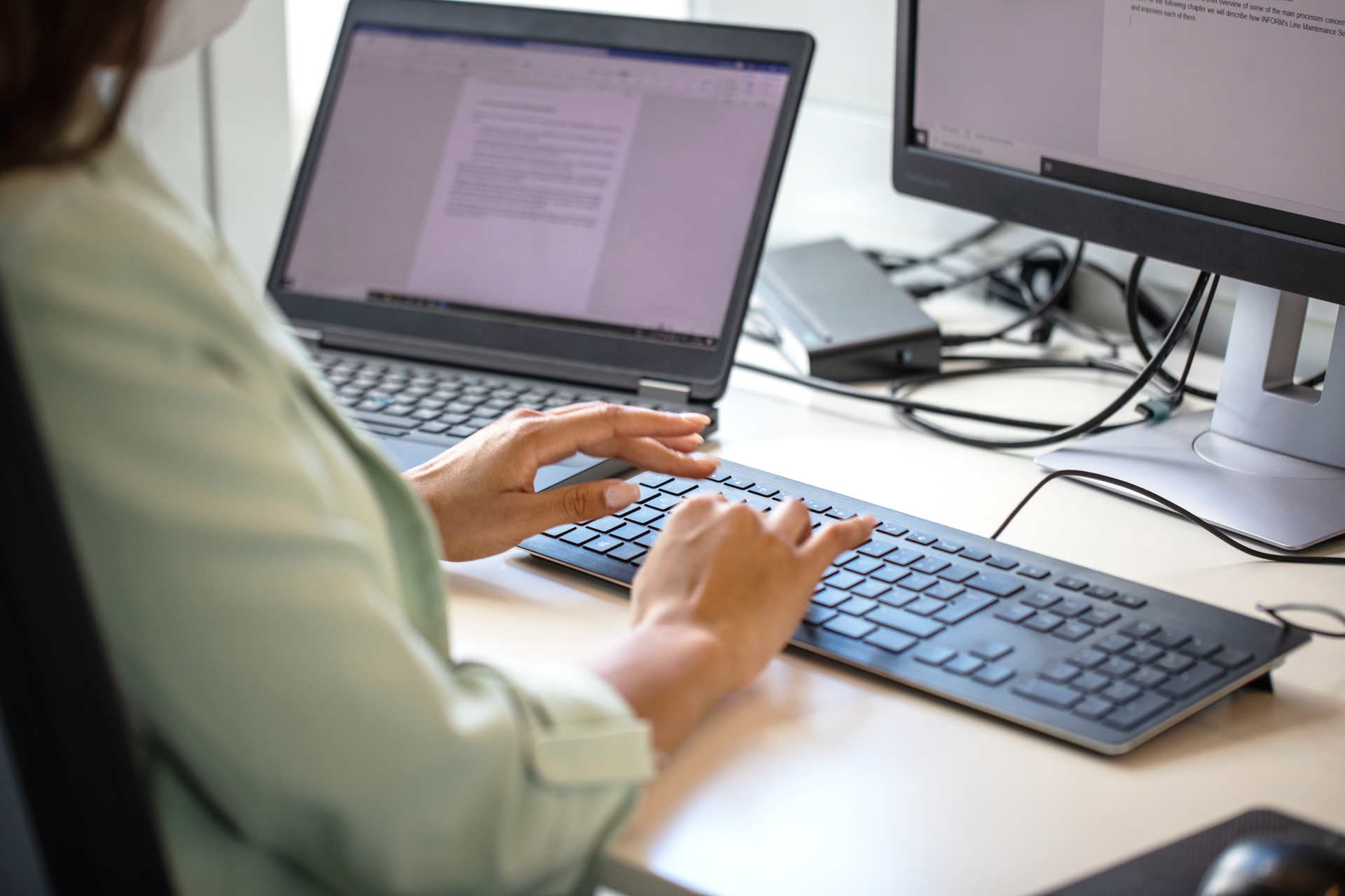 Eine Person tippt an einer Tastatur, während zwei Computerbildschirme auf einem Schreibtisch sichtbar sind, einer mit einem geöffneten Textdokument.