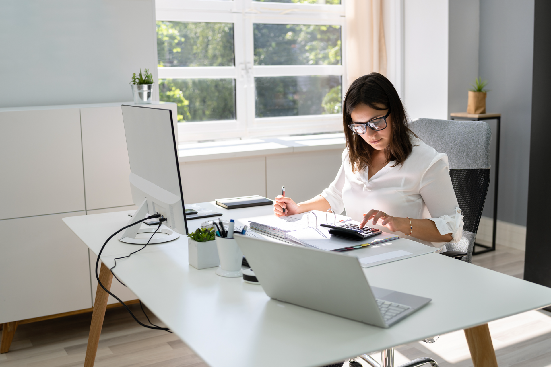 Eine Frau sitzt an einem modernen Schreibtisch in einem hellen Büro, während sie mit einem Taschenrechner arbeitet und Notizen auf einem Notizblock macht. Auf dem Tisch stehen mehrere Büromaterialien, ein Computerbildschirm und eine Laptop, während Sonnenlicht durch das Fenster strömt.