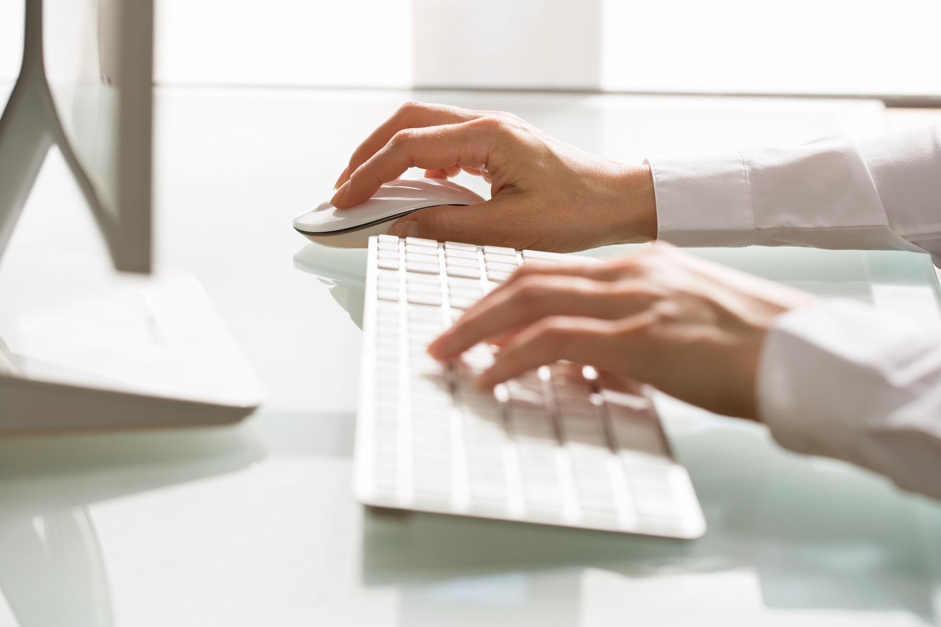 Eine Hand verwendet eine Computermaus, während die andere Hand auf einer weißen Tastatur tippt. Im Hintergrund ist ein Bildschirm auf einem Tisch zu sehen.