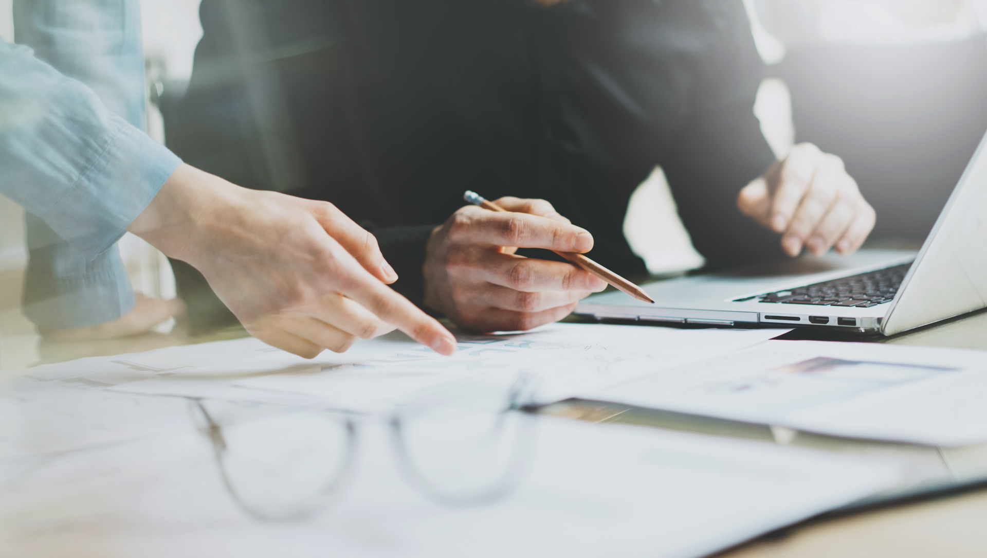 Zwei Personen arbeiten gemeinsam an einem Tisch, während eine Hand auf ein Papier zeigt und die andere einen Stift hält; ein Laptop ist geöffnet und leicht sichtbar, sowie Brille und weitere Papiere liegen davor.